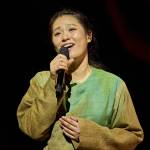 Singer (Ai Ninomiya) in a green shirt and singing into a microphone.